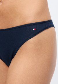 Detailní záběr na osobu v námořnicky modrých žebrovaných spodních dílech bikin s malým červenobíločerveným logem na boku.