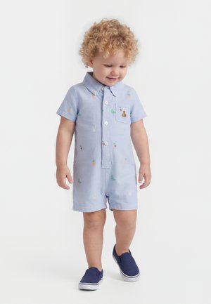 Batole s kučeravými vlasmi v svetlom modrom krátkym overal s motívmi ovocia a zajka a v námorníckych slip-on topánkach, ktoré sa pozerá nadol.