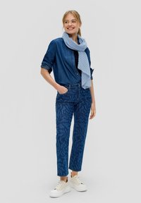 Chemise en denim avec des manches retroussées, jeans bleus à motifs et écharpe bleu clair. Des baskets plates blanches complètent la tenue.