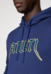 Mand iført en marineblå hættebluse med et gult og blåt stiliseret "ATHLET1" logo på brystet og snøringer hængende ned.
