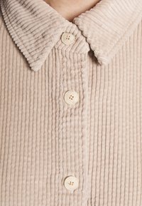 Camicia di velluto a coste color beige, con una texture rigata, caratterizzata da un colletto aperto e quattro bottoni circolari. La vista ravvicinata mostra il motivo delle righe verticali.