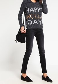 Grijze trui met "HAPPY HOLLA DAY" in lichtgrijs en gouden pailletten. Gecombineerd met zwarte slim fit jeans en zwarte instappers. Zwarte tas.