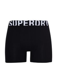 Bañadores boxer negros hechos de un tejido elástico, con una ancha cinturilla elástica blanca que lleva el texto "SUPERDRY" en letras grandes.