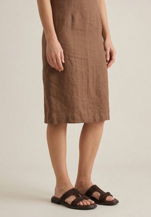 Vrouw draagt een knielange bruine linnen jurk en donkerbruine open teenslippers staand op een lichte achtergrond.