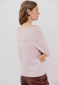 Světle růžový svetr přes ramena z jemného materiálu, se žebrovaným výstřihem a manžetami, v kombinaci s hnědými koženými kalhotami.