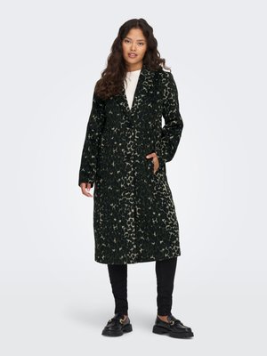 Jeune femme portant un long manteau léopard vert foncé et noir, pantalon noir, mocassins noirs, debout avec une main dans la poche.