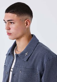 Giacca grigia con trama a cialda, collo con risvolto, tasca sul petto ben visibile e dettaglio a stella d'argento sulla revers, indossata sopra una camicia bianca.