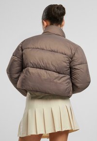 Smeđa puf jakna s visokim ovratnikom i tkaninom od kvadrata. Model nosi plisiranu bež suknju ispod.
