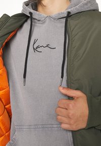 Homme portant un sweat à capuche gris avec des cordons noirs et un logo, partiellement couvert par une veste verte avec une doublure matelassée orange, tenant la veste ouverte.