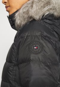 Chaqueta acolchada negra con capucha forrada de piel sintética gris. Parche con logo en la manga con diseño redondo y texto. Material liso y aislante.