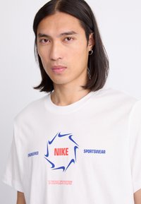 Balta medvilnės marškinėliai su apvalia mėlyna ir raudona grafika, kurios centre yra "NIKE", o šonuose rašoma "ENGINEERED" ir "SPORTSWEAR".