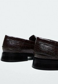 Mørkebrune læder loafers med krokodille tekstur, lave stablede sorte hæle, placeret side om side på en ensfarvet lys baggrund.