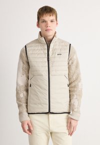 Tânăr îmbrăcat cu o vestă fără mâneci, bej, matlasată, cu dantelă neagră, peste un pulover bej cu model și pantaloni de culoare deschisă, pe un fundal alb.