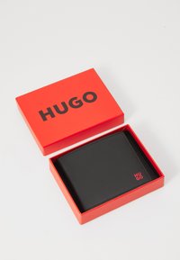 Schwarzes Lederportemonnaie in einer roten Box mit "HUGO" in fetten schwarzen Buchstaben aufgedruckt. Das Portemonnaie verfügt über eine gestickte Kante und ein kleines Logodetail.