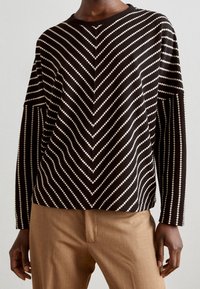 Maglione a righe marroni e crema con motivo a zigzag, scollatura rotonda e maniche lunghe, abbinato a pantaloni beige.