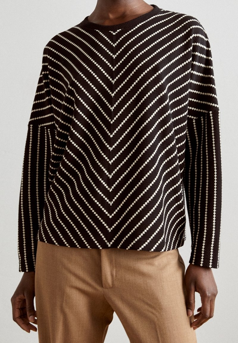 Maglione a righe marroni e crema con motivo a zigzag, scollatura rotonda e maniche lunghe, abbinato a pantaloni beige.