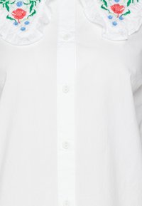 Camicia bianca con colletto arricciato, decorata con ricami floreali colorati in rosso, blu e verde, realizzata in tessuto morbido.