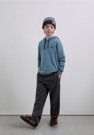 Sudadera con capucha azul claro con cordones azul oscuro y pequeño logo, combinada con pantalones de pana gris oscuro y zapatos marrones.