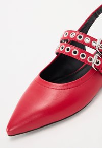 Ballerine in pelle rossa a punta con doppie cinghie che presentano occhielli metallici e fibbie, caratterizzate da una texture liscia e un design minimalista.