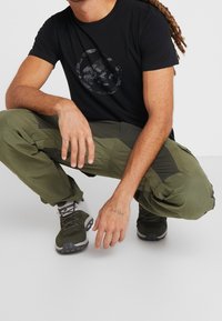 Camiseta negra de cuello redondo con diseño reflectante, pantalones cargo verdes oliva con múltiples bolsillos y botas de senderismo verde oscuro.