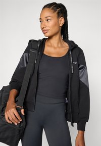 Felpa nera con zip e pannelli laterali, abbinata a una canotta nera e leggings. La modella tiene una borsa da palestra nera con dettagli texture.