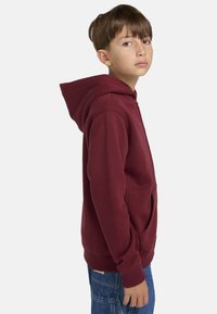 Sudadera con capucha burdeos con un bolsillo delantero, puños acanalados y un diseño limpio. Combinada con jeans de mezclilla azules, vista de perfil lateral.