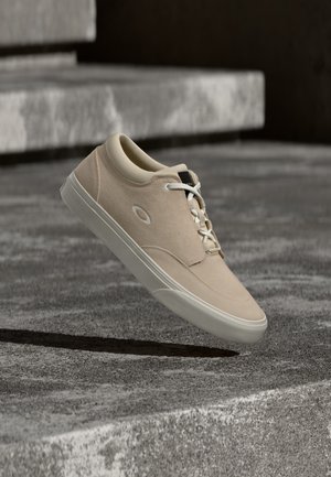 Beige casual sneaker met witte veters zwevend boven getextureerde betonnen trappen in een schemerig verlichte stedelijke omgeving.