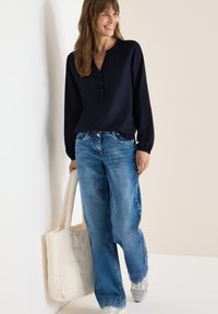 Blouse bleu marine à manches longues avec une patte de boutonnage, associée à un jean bleu ample. Le modèle porte un tote matelassé blanc et des baskets argentées.