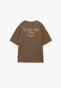 Brun bomulds t-shirt med løs pasform, der har hvid tekst på ryggen: "Le Petit Cafe Paris" samt adresseoplysninger i en afslappet skrifttype.