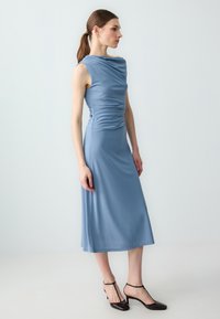 Abito midi blu con scollatura drappeggiata, silhouette aderente e texture liscia. Indossato con scarpe con tacco scuro.