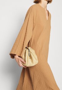 Femme portant une robe ample texturée couleur camel, tenant une petite pochette tissée beige avec un fermoir métallique sur fond uni.