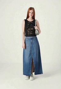 Νεαρή γυναίκα με ίσια κόκκινα μαλλιά, ντυμένη με μαύρη αμάνικη μπλούζα με δαντελένιες λεπτομέρειες, μακριά τζιν φούστα σε μπλε απόχρωση με μπροστινή σχισμή, λευκά sneakers και ασημένια τσάντα.