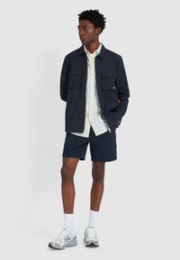 Marineblauer Zip-Jacke mit zwei Fronttaschen, kurzärmeliges gestreiftes Shirt, marineblaue strukturierte Shorts und silberne Sportschuhe mit weißen Socken.