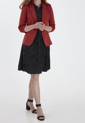 Frau trägt einen rostfarbenen Blazer über einem schwarzen, geblümten Kleid und schwarze Sandalen mit Blockabsatz und Knöchelriemen, steht mit gefalteten Händen.