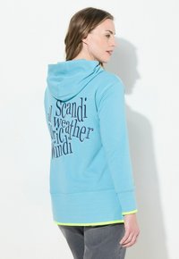 Felpa con cappuccio di colore azzurro chiaro, caratterizzata da un design grafico di testo in blu scuro sul retro, con dettagli di colore giallo neon lungo l'orlo e i polsini.