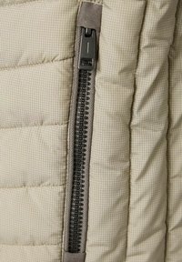 Beige gesteppter Stoff mit einem grauen Reißverschluss und einem schwarzen Zugtab, vor einem karierten Muster. Strukturiere Oberfläche mit sichtbaren Nähten.