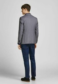 Grå blazer med almindelig lapel, enkelt rygslids og sorte knapper, parret med mørke denimjeans og sorte sko. Tekstureret stof synligt.