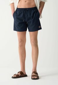 Pantaloni da bagno blu navy con vita elastica, cordino e tasche laterali. Abbinati a sandali marroni con larghe cinghie.