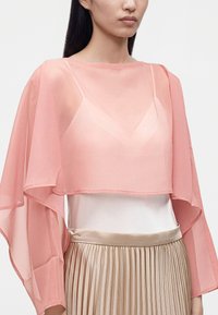 Femme portant un petit cape rose transparent par-dessus un haut blanc et une jupe beige plissée, face à l'avant sans expression faciale visible.