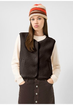 Jeune femme portant un bonnet en tricot rayé, un pull crème à manches longues, une jupe marron foncé boutonnée et un gilet noir texturé, les mains dans les poches.