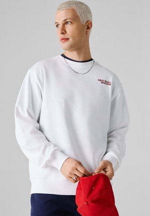 Valge pika varrukaga ja ümar kaelusega dressipluus, mille rinnal on väike punane "RED BULL RACING" logo, mida hoiab punane nokamüts.