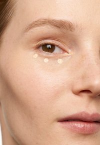 Concealerprickar i en beige nyans applicerade under ögat på slät, ljus hy med jämn struktur och minimala fläckar. Naturligt ljus reflekteras.