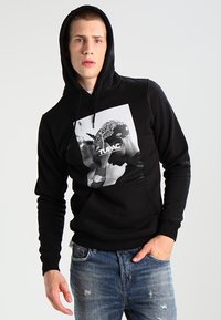 Schwarzer Hoodie aus weichem Material mit einem großen Grafikdruck von Tupac. Er hat eine Fronttasche und wird mit ausgewaschenen Jeans kombiniert.