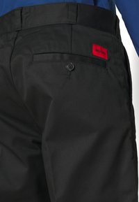 Svarta byxor med en bakficka med knapp. En röd tygpatch med "HUGO" syns på baksidan. Slät textur och strukturerad design.