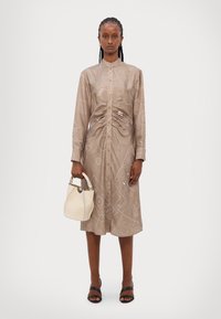 Beige košeľové šaty s bielymi geometrickými vzormi, s naťahovacím pásom. Modelka drží krémovú kabelku a má na sebe čierne sandále na ramienka.