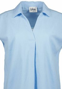 Camicia azzurro chiaro con colletto a V, maniche corte e fronte plissettato. L'etichetta riporta "blu" e "BLUE SEVEN" con taglia 36.