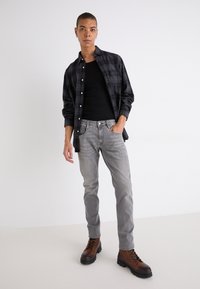 Svart rutig skjorta över en svart linne, grå slim-fit jeans och bruna snörkängor med tjock sula. Modellen står med en hand i fickan.