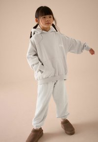 Grå oversized hoodie med framficka, med en liten vit logotyp. Matchad med ljusgrå sweatpants och bruna slip-on skor.