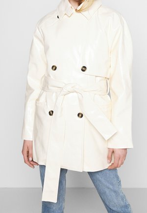 Femme portant un trench-coat crème ceinturé avec de gros boutons et un jean bleu, debout devant un fond clair uni.