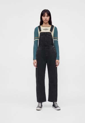 Jeune femme portant un pull rayé bleu canard et crème sous une salopette en jean noire avec des baskets montantes noires, debout.